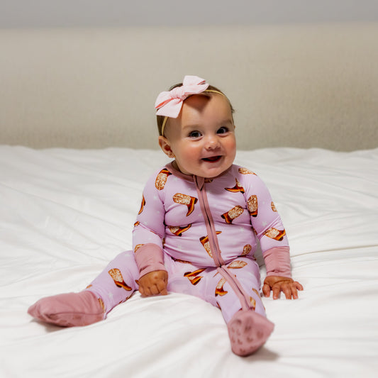 Baby wearing a pink onesie with animal prints sitting on a bed.