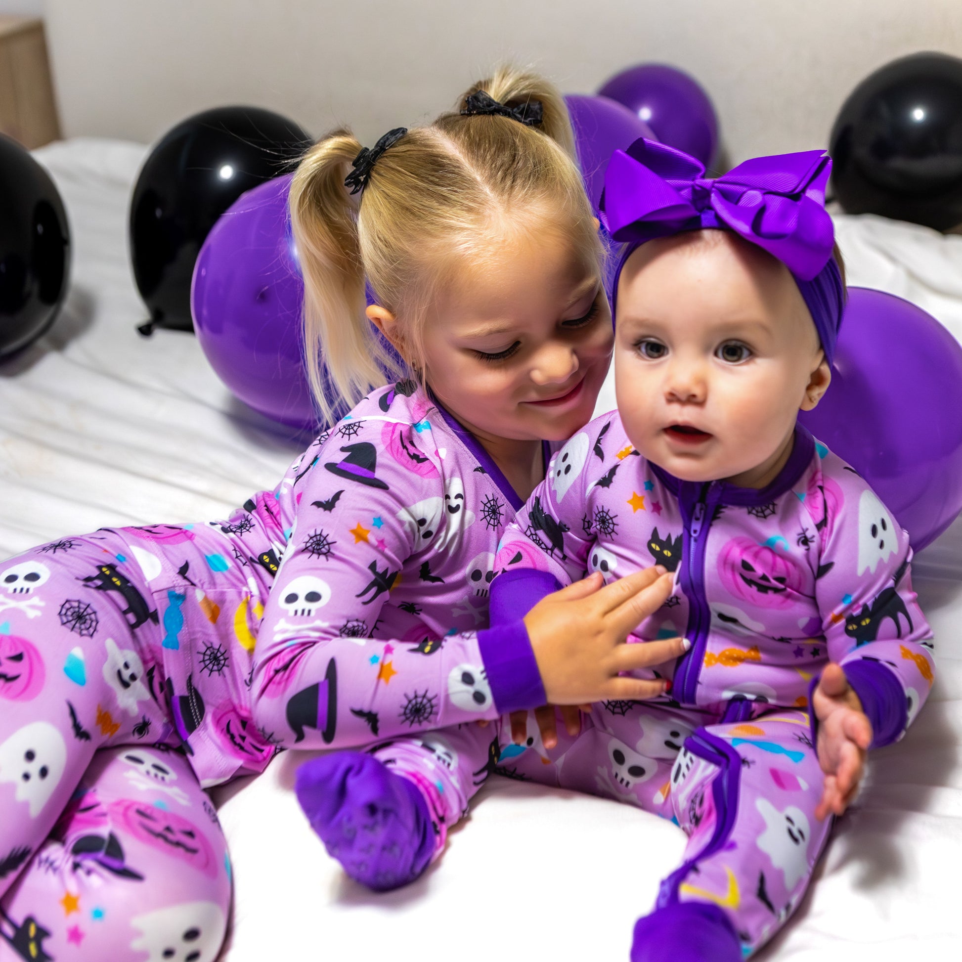 Two children in Halloween-themed pajamas surrounded by balloons on a bed.